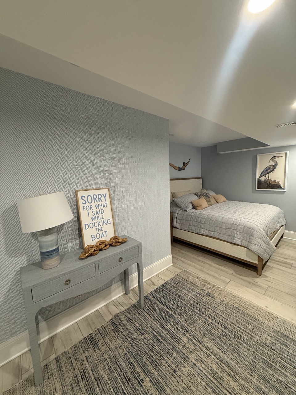 Lower level guest bedroom with light blue textured walls, a gray console table with a lamp, a bed and wood look tile floor