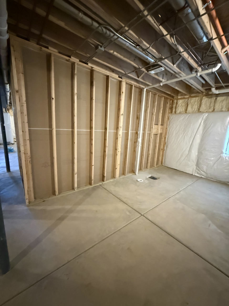 Basement bedroom walls framed in wood studs on a concrete floor, with blanket insulation on the foundation wall and ducts and pipes open overhead