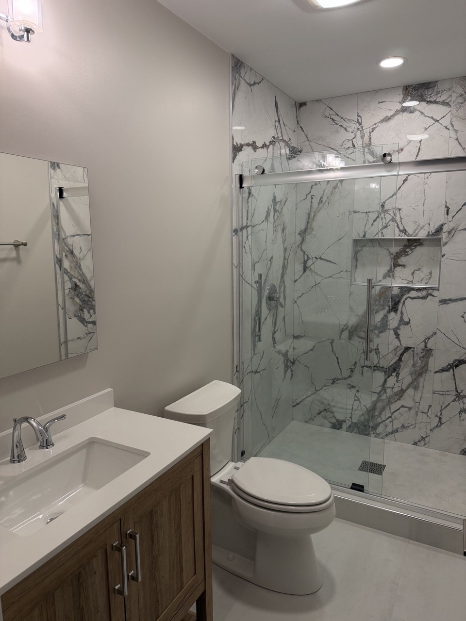 Bathroom with a marble look tile shower, a sliding glass door, a built in niche, a wood vanity with a white top and a toilet