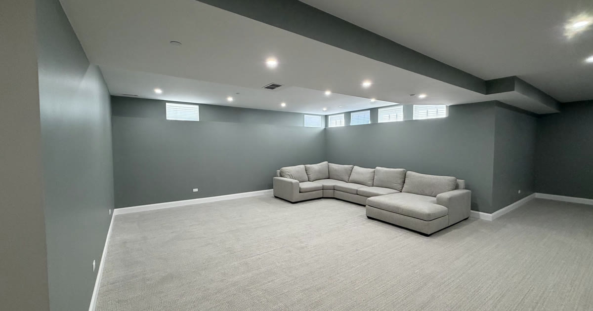 Finished basement family room with gray walls, light carpet, recessed lights, a soffit boxing in the ducts and a row of small windows near the ceiling