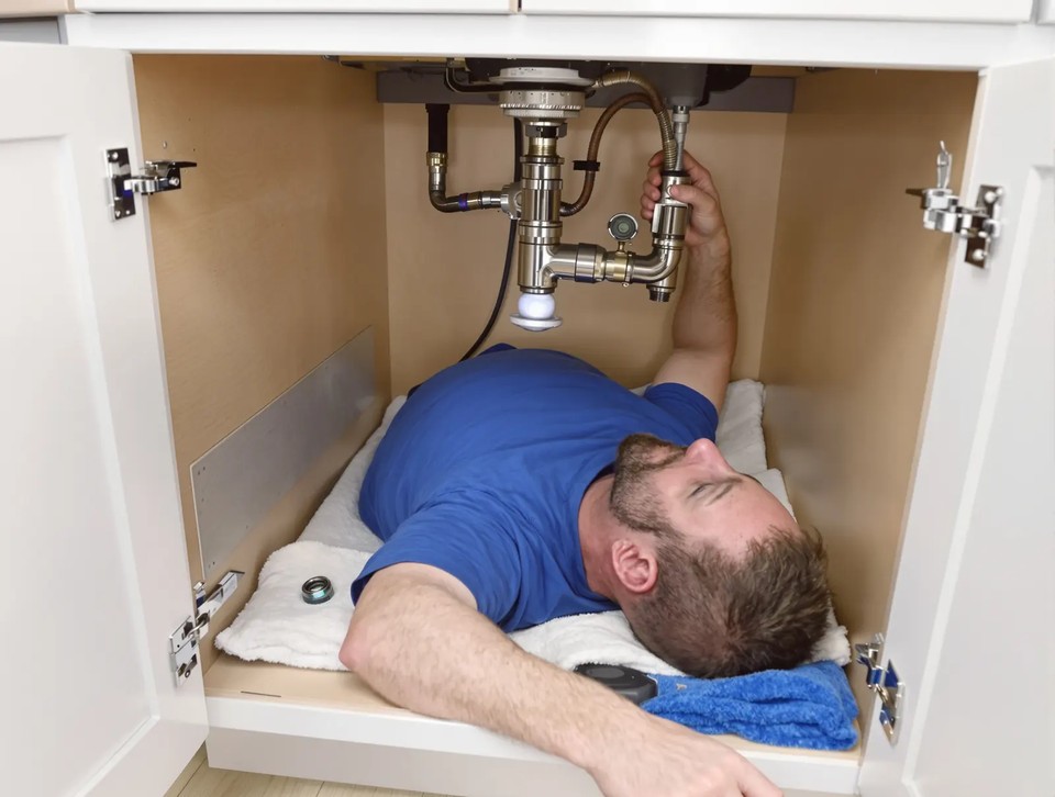 Handyman lying inside an open kitchen sink cabinet on a towel, reaching up to the faucet supply lines and drain pipes