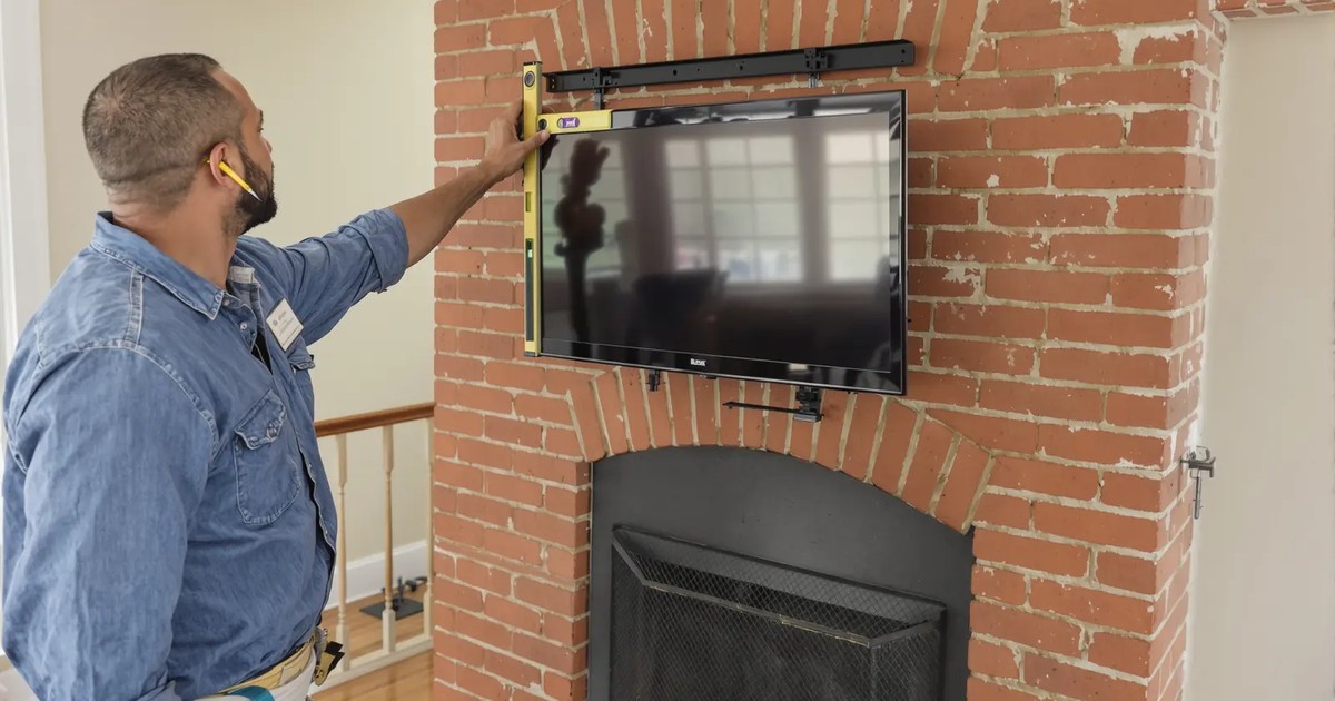 Handyman in a denim shirt leveling a TV mount on a brick fireplace wall, with a drill and tools on a drop cloth below